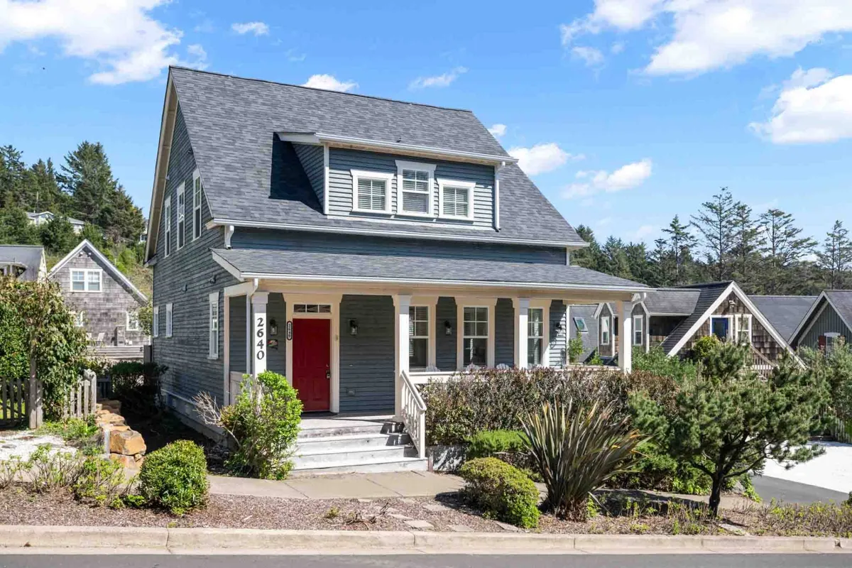 The inviting front porch of a home, featuring a vibrant red door, lush landscaped garden, and sunny ambiance. The space is designed for gatherings, outdoor relaxation, and easy transitions from inside comforts to outdoor fun.