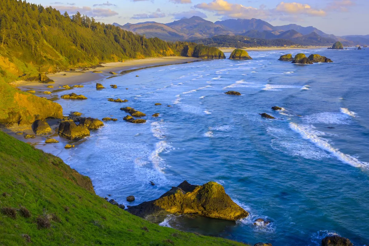 A breathtaking view of the rocky coastline framed by gentle waves, sandy beach, and distant mountains. The natural scenery provides a sense of place and adventure, inviting exploration and appreciation of Lincoln City's coastal beauty.