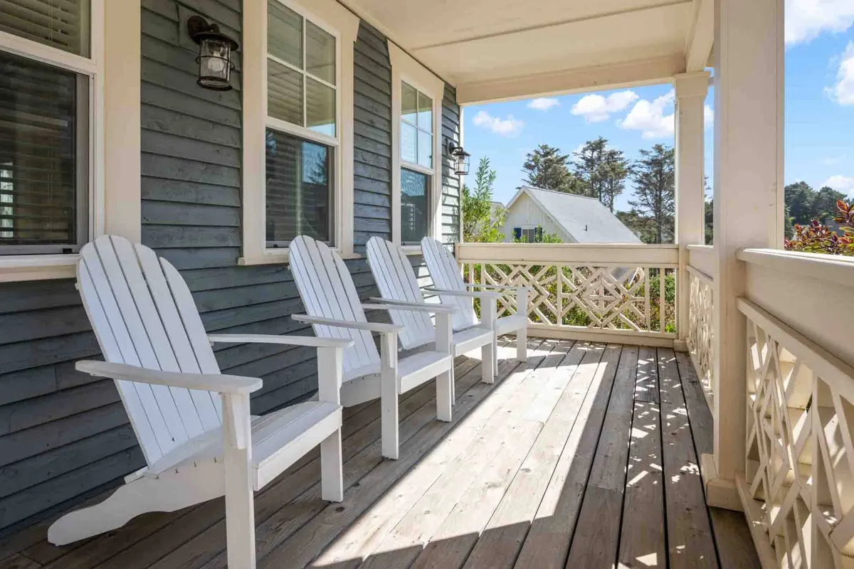 Four white Adirondack chairs are arranged on a wooden deck with a decorative railing, providing a comfortable space to relax, read, or enjoy the view. The atmosphere is calm and peaceful, ideal for enjoying ocean breezes or morning coffee.