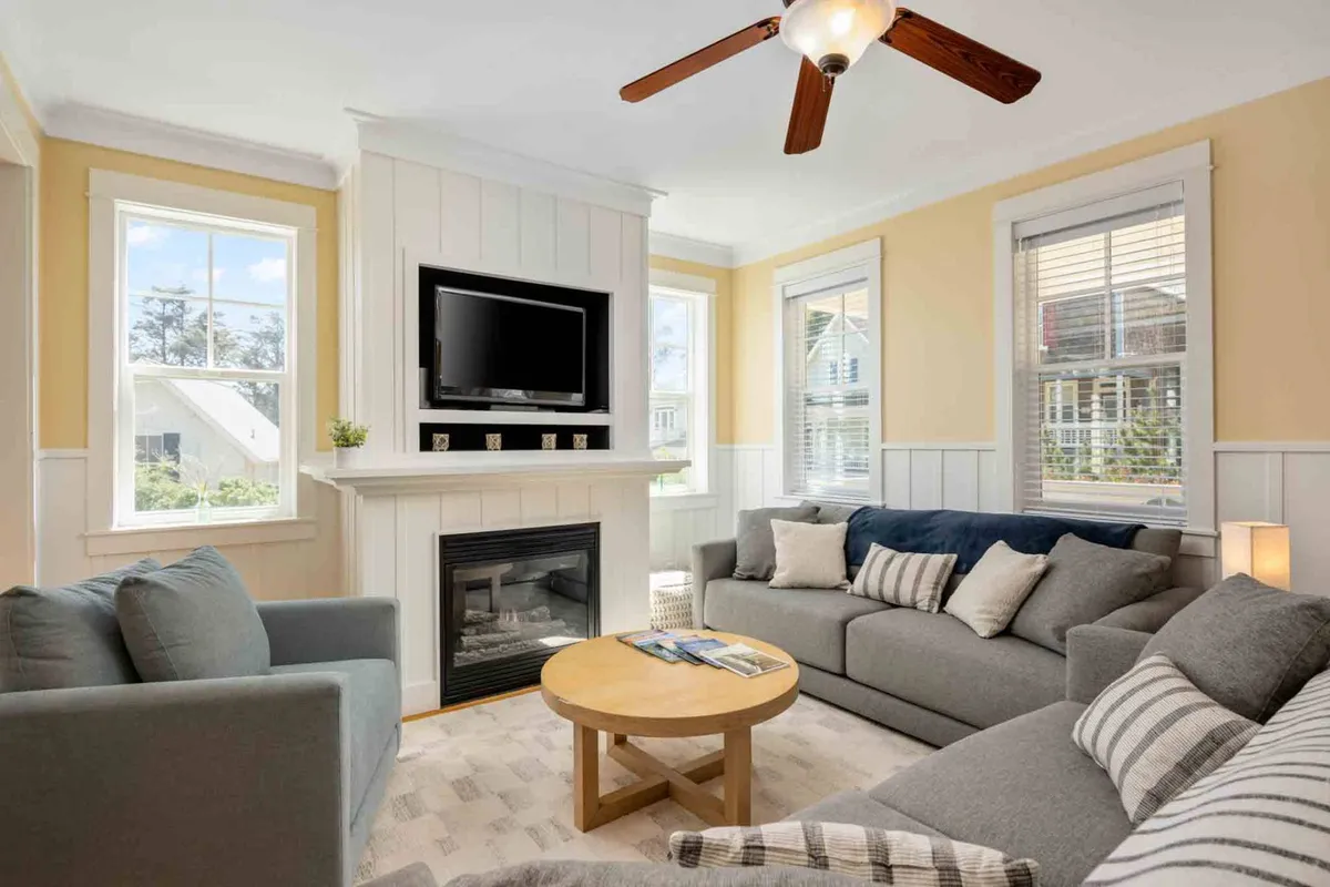 A warm, cozy living room with a glowing fireplace, comfortable sofas arranged for conversation, a coffee table with puzzles, and large windows inviting in natural light. The setting encourages relaxation and indoor comfort, especially on rainy or chilly days.
