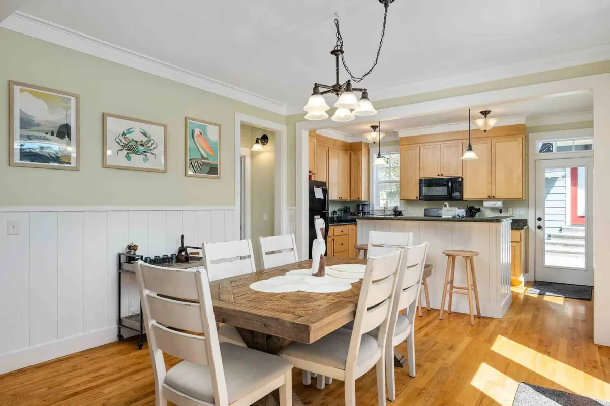 This interior scene captures the inviting dining area adjacent to the kitchen, featuring a wooden dining table, stylish light fixture, kitchen area in the background, and wall art. The space exudes warmth, encouraging family meals and shared moments.