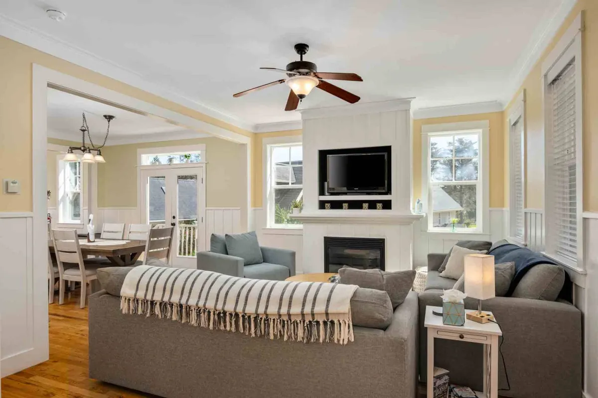 A welcoming open-concept living and dining area bathed in natural coastal light. Comfortable sofas face a cozy fireplace, with a dining table visible in the background. Sunlight pours in through large windows, creating an inviting space perfect for both relaxation and lively gatherings.