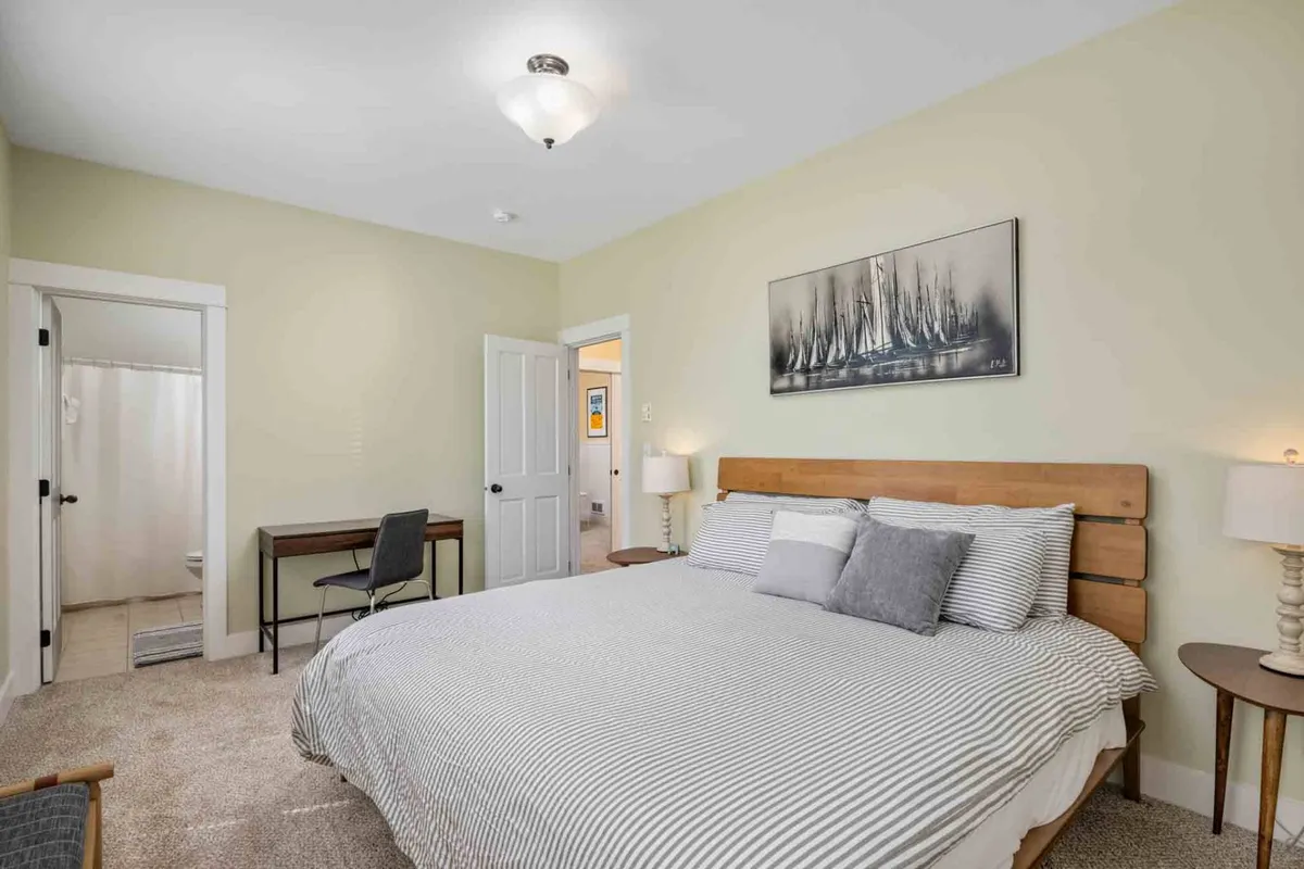 A spacious bedroom featuring a large bed with striped bedding, a desk and chair setup perfect for quiet work or reading, and framed artwork on the wall. The space is bathed in natural light, creating a serene and welcoming atmosphere.