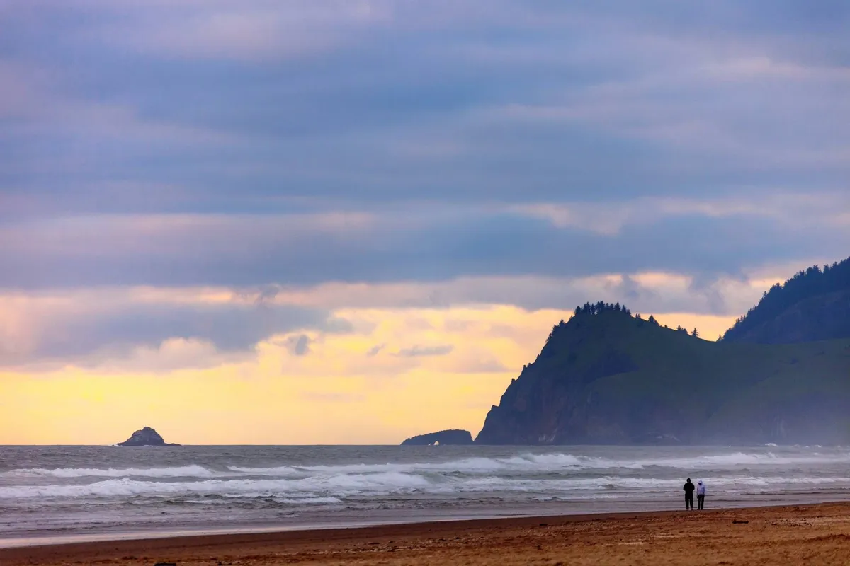 A couple strolls along a sandy beach with ocean waves and distant cliffs in the background, embodying the coastal proximity and opportunities for local exploration. The mood is peaceful and romantic, echoing the experiences available just a walk away from the retreat.