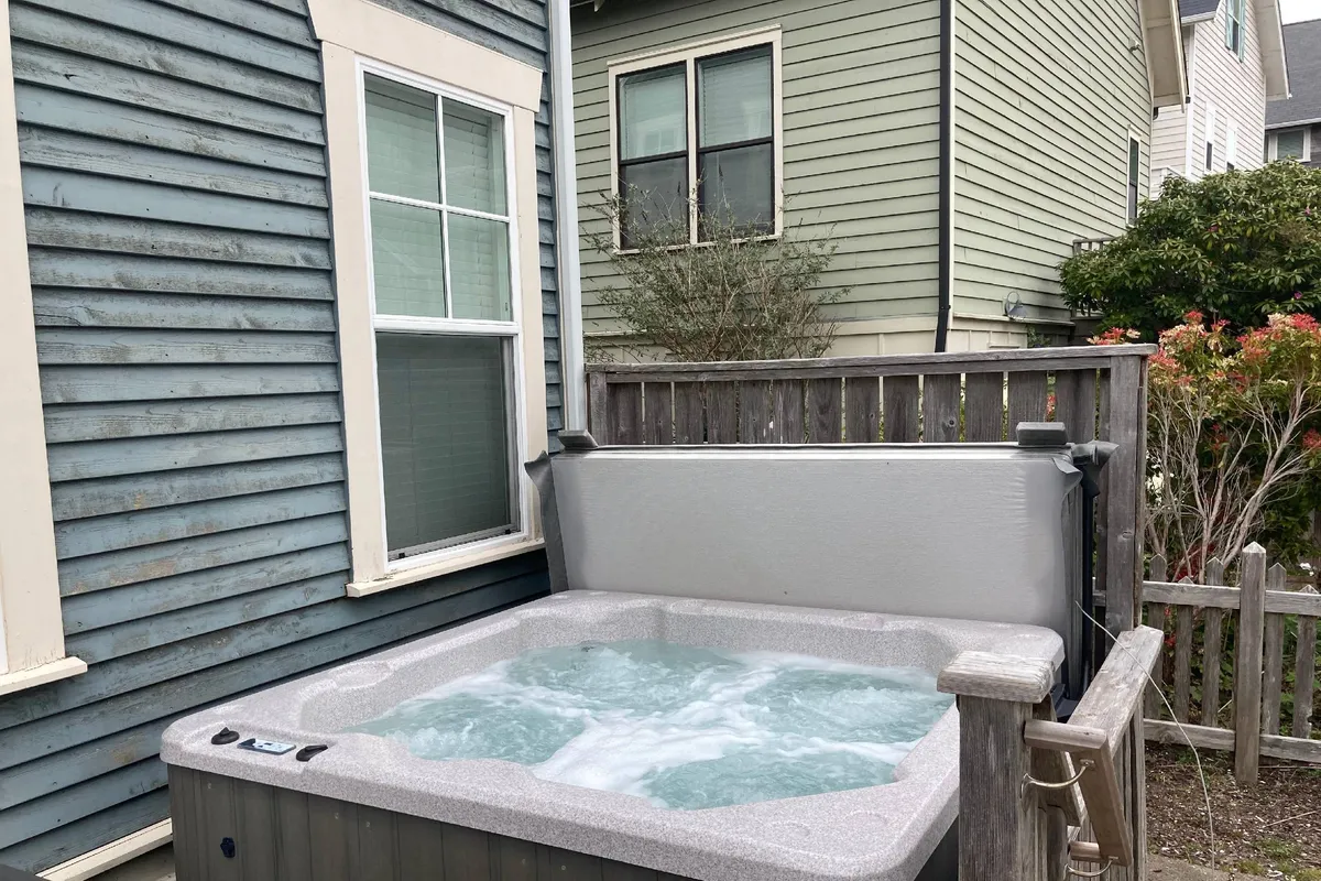 This exterior shot features a private hot tub surrounded by a wooden fence, with neighboring buildings visible in the background. The area exudes a sense of relaxation and privacy, perfect for unwinding after a day out in Lincoln City.