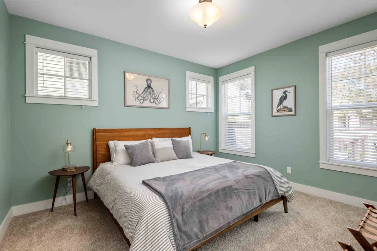 A tranquil bedroom featuring a comfortable bed dressed in gray and white bedding, whimsical octopus and bird artwork, a wooden bedside table, and a soft-glow table lamp. The room feels relaxing and private—perfect for a quiet retreat.