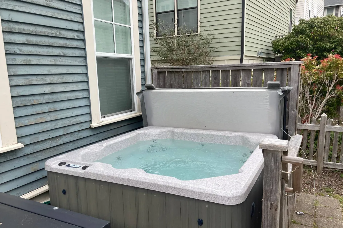 A private outdoor hot tub sits behind a wooden fence, with windows of the house visible in the background. The area is designed for relaxation and socializing, offering a peaceful and inviting spot for soaking up the coastal ambiance.