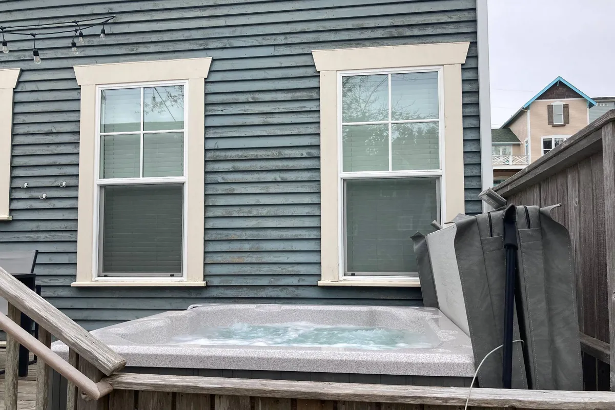 An inviting exterior view features a bubbling hot tub on a private deck, bordered by a wooden fence and accessible via stairs. The space is designed for relaxation and socializing, with the home’s windows in the background creating a seamless indoor-outdoor connection.
